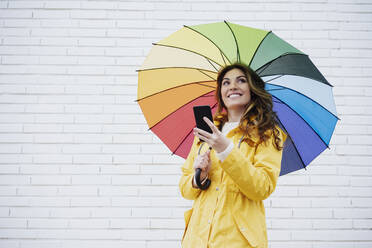 Frau hält Smartphone und Regenschirm vor einer Backsteinmauer - EBBF05160