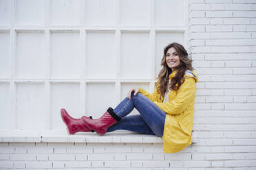 Young woman with raincoat and rubber boots sitting on brick wall - EBBF05162