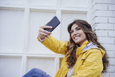 Lächelnde junge Frau nimmt Selfie durch Smartphone an der Wand - EBBF05165