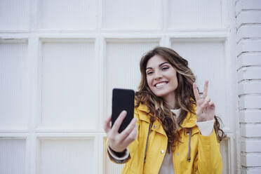 Frau zeigt Friedenszeichen und macht ein Selfie mit ihrem Smartphone vor einer Mauer - EBBF05166