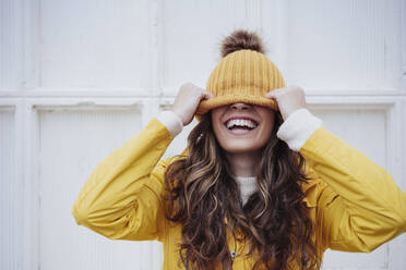 Playful woman covering face with knit hat in front of wall - EBBF05171