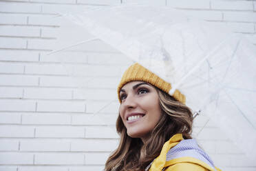 Lächelnde Frau mit transparentem Regenschirm an der Wand - EBBF05173
