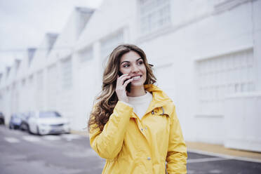 Lächelnde Frau in gelbem Regenmantel, die mit einem Mobiltelefon spricht - EBBF05181