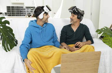 Smiling gay couple wearing VR glasses looking at each other sitting on sofa - JCCMF04868