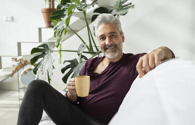 Smiling man holding coffee cup on sofa - JCCMF04870
