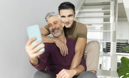 Lächelnder Mann nimmt Selfie mit Ehemann durch Handy zu Hause - JCCMF04889