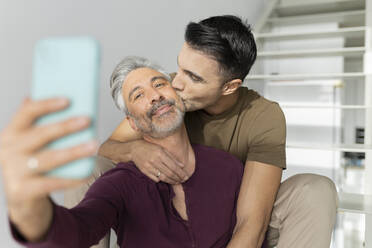 Glücklicher schwuler Mann, der ein Selfie mit seinem Mann macht, der ihn zu Hause auf die Wange küsst - JCCMF04890