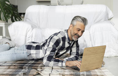 Lächelnder Mann mit Laptop im Wohnzimmer liegend - JCCMF04900