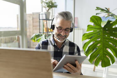 Smiling businessman with headphones and laptop using tablet PC at home - JCCMF04914