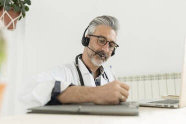 Arzt mit Kopfhörern beim Schreiben auf einem Klemmbrett im Home Office - JCCMF04923