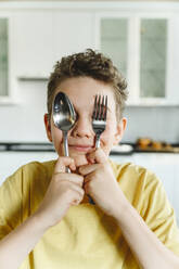 Smiling boy covering eyes with fork and spoon at home - EYAF01857