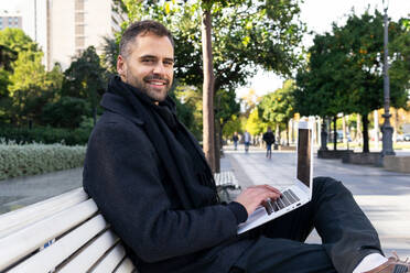 Seitenansicht eines nachdenklichen männlichen Unternehmers in Freizeitkleidung, der auf einer Holzbank sitzt und an einem Projekt auf einem Laptop auf einer Straße am Tag arbeitet - ADSF32952