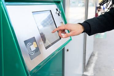 Abgeschnittene unerkennbare männliche Person in schwarzem Mantel steht mit der Hand in der Tasche und kauft eine Fahrkarte auf dem Bahnhof in der Stadt bei Tag - ADSF32953