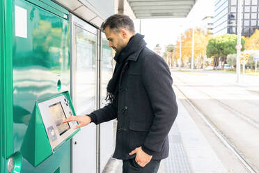 Side view of serious male in black coat standing with hand in pocket and buying ticket on railway station in city in daytime - ADSF32954