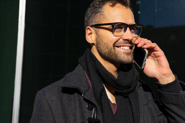 Cheerful male in eyeglasses and coat talking on smartphone and looking away on street in sunlight - ADSF32957