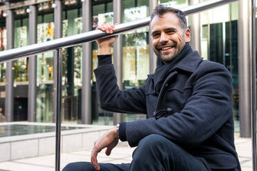 Lächelnder Mann in schwarzem Mantel, der auf einer Treppe in der Nähe eines Metallgeländers auf der Straße sitzt und in die Kamera blickt, gegen ein modernes Gebäude in der Tageszeit - ADSF32961