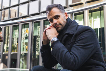 Male in black coat sitting on stairs near metal railing on street and looking at camera against contemporary building in daytime - ADSF32962