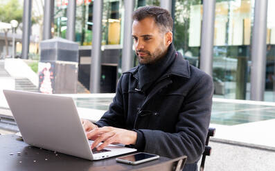 Lächelnder Geschäftsmann in schwarzem Mantel, der an einem Tisch in einem Café sitzt und auf einem Laptop surft - ADSF32963
