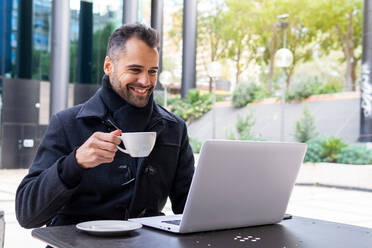 Smiling businessman in black coat drinking cup of tea and talking on video call via laptop while sitting at table in cafe in daytime - ADSF32965