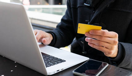 Crop unrecognizable male entrepreneur in black coat looking at details of credit card in hands while sitting at table with laptop on terrace of cafe in daytime - ADSF32966