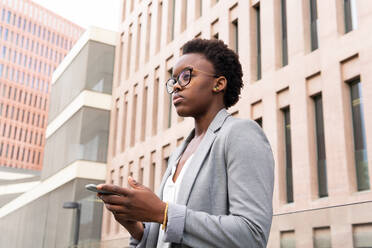 Seitenansicht einer schwarzen Frau in hellgrauer Jacke und mit Brille, die ein Mobiltelefon benutzt, während sie auf der Straße in der Nähe eines modernen Bürogebäudes steht - ADSF32967