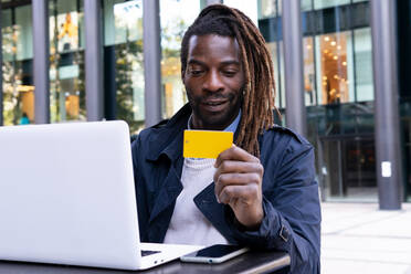 Positiver afroamerikanischer junger Mann, der eine Bestellung in einem Internetshop mit Kreditkarte auf einem Laptop bezahlt, der an einem Tisch mit Smartphone auf der Terrasse eines Einkaufszentrums sitzt - ADSF32973