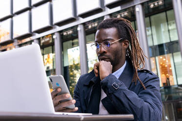 Gelassener afroamerikanischer junger Mann, der an einem Tisch mit einem Laptop auf der Terrasse eines Einkaufszentrums sitzt und mit seinem Smartphone Nachrichten schreibt - ADSF32974