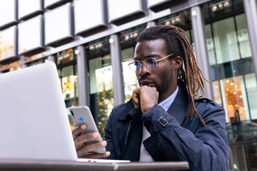 Serene African American young man messaging on smartphone sitting at table with a laptop on terrace of shopping center - ADSF32974