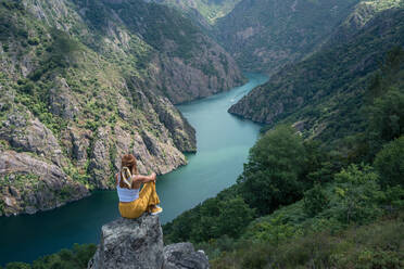 Rückenansicht einer anonymen Reisenden, die am Rande eines felsigen Berges sitzt und die herrliche Landschaft des Flusses Syl Canyon in Ribeira Sacra in Galicien Spanien bewundert - ADSF32976