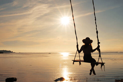 Rückansicht der Silhouette einer anonymen jungen Frau mit Hut, die auf einer Schaukel sitzt und den herrlichen Sonnenuntergang in Cambados in Spanien genießt - ADSF32977