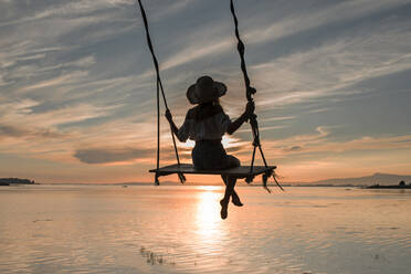 Rückansicht der Silhouette einer anonymen jungen Frau mit Hut, die auf einer Schaukel sitzt und den herrlichen Sonnenuntergang in Cambados in Spanien genießt - ADSF32978