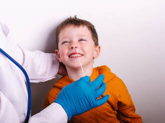 Von oben: ein Arzt in Uniform und Handschuh, der einen COVID 19-Abstrichtest durchführt, und ein fröhlicher Junge mit einem breiten Grinsen an der grauen Wand - ADSF32983