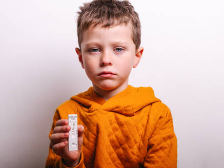 Ill boy in orange hoodie demonstrating positive COVID 19 antigen test against gray background - ADSF32984