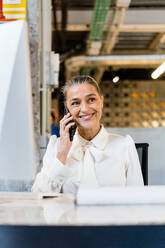 Lächelnder weiblicher Manager in elegantem Outfit, der wegschaut, während er während der Arbeit in einem modernen hellen Büro am Schreibtisch telefoniert - ADSF32993
