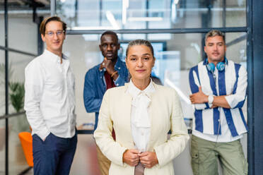 Weibliche Angestellte in formeller Kleidung schaut in die Kamera, während sie in der Nähe einer Gruppe von männlichen Kollegen in einem hellen, modernen Büro steht - ADSF32998