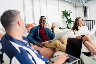 Gruppe positiver multiethnischer Mitarbeiter mit Netbooks, die auf Sackstühlen sitzen, während sie gemeinsam an einem Geschäftsprojekt in einem modernen Büro arbeiten - ADSF33000