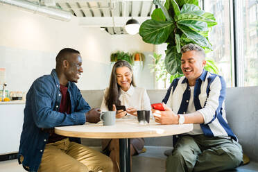 Gruppe positiver multirassischer Mitarbeiter, die mit ihren Handys surfen, während sie während der Pause in einem modernen hellen Café an einem Tisch mit heißen Getränken sitzen - ADSF33001