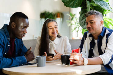 Gruppe positiver multirassischer Mitarbeiter, die mit ihren Handys surfen, während sie während der Pause in einem modernen hellen Café an einem Tisch mit heißen Getränken sitzen - ADSF33002