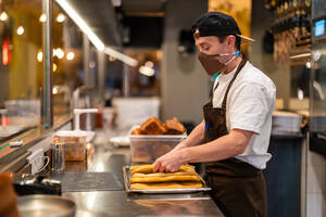 Side view of male cook in protective mask standing at stainless counter with steel tray of baked flatbread in restaurant open kitchen during pandemic - ADSF33004