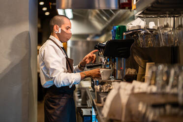 Seitenansicht eines Kellners in Uniform und Schutzmaske, der während der Coronavirus-Pandemie in einem Restaurant die Knöpfe einer Kaffeemaschine drückt und ein Getränk aufbrüht - ADSF33005