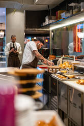Male cook preparing food at counter in modern open kitchen near waiter using cellphone in restaurant during work at COVID pandemic - ADSF33009
