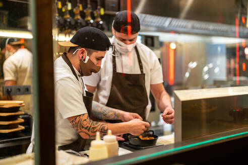 Männliche Köche in eleganter Uniform und mit Schutzmasken zur Verhinderung der Ausbreitung von COVID 19 bei der Zubereitung von Mahlzeiten in der Küche eines Restaurants durch Glas - ADSF33010