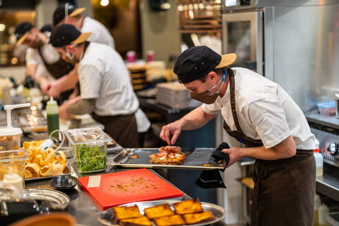 Männlicher Koch, der eine Füllung auf ein Fladenbrot auf einem rostfreien Tablett aufträgt, mit Schutzmaske und Uniform in einem Restaurant - ADSF33012