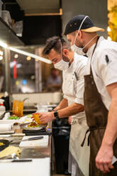 Side view of professional chef in mask serving plate with delicious dish with colleague in restaurant kitchen during coronavirus pandemic - ADSF33013