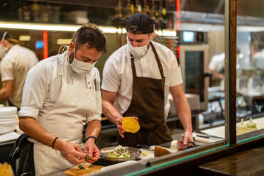 Through glass of professional male chef serving delicious meal on plate with assistant helping in cooking process in restaurant during coronavirus pandemic - ADSF33014