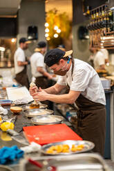 Side view of professional male cook in protective mask adding cream from pastry bag while preparing delicious burger in restaurant kitchen at work with colleagues during coronavirus epidemic - ADSF33016