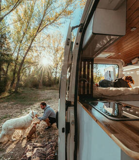 Man caressing Staffordshire Terrier and White Swiss Shepherd dog while sitting near camper van with wife in Cuenca green forest - ADSF33021