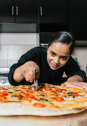 Fokussierte junge ethnische weibliche Köchin in Uniform schneidet frisch zubereitete Pizza mit Messerwalze - ADSF33026