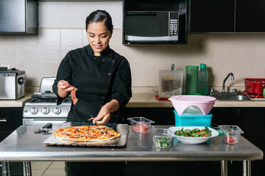 Junger ethnischer weiblicher Koch in Uniform, der Schinken auf eine leckere Pizza gibt, während er auf der Küchentheke eines modernen Cafés kocht - ADSF33027