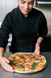 Junge positive Köchin Frau in schwarzer Uniform von leckeren frisch gekochten Pizza auf den Tisch gelegt - ADSF33028